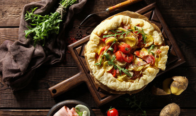 Concept of French and Italian cuisine. Pie with vegetables and bacon. Potatoes, cherry tomatoes, onions, bacon, arugula and spices on an old wooden background. Background image, copy space