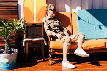 Stylish portrait of a young woman on an orange sofa outdoors in the interior of the terrace of a trendy lounge cafe. Summer fashion concept.