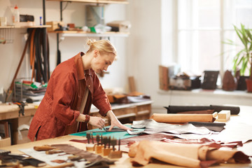 Horizontal side view portrait of modern female artisan doing leather craftwork standing at table in her workshop