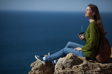 Fototapeta na wymiar Beautiful young woman traveler with a backpack drinking morning coffee and looking at the sea. Copy space. The concept of freedom , morning coffee, travel and an active lifestyle.
