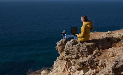Fototapeta na wymiar A freelancer girl is working typing on a laptop and looking at a monitor with a beautiful view of the open air sea sky. Traveling with a computer. Online dream job.