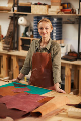 Vertical medium long portrait of beautiful young woman wearing apron standing at work table with leather material on it looking at camera smiling