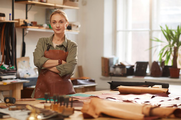 Horizontal portrait of confident Caucasian woman wearing apron standing with arms crossed in modern leather craft workshop looking at camera, copy space