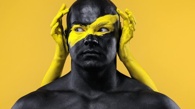 Man with female hands on the body. Bodybuilder athlete with yellow face art and black body paint. Colorful portrait of the guy with bodyart.
