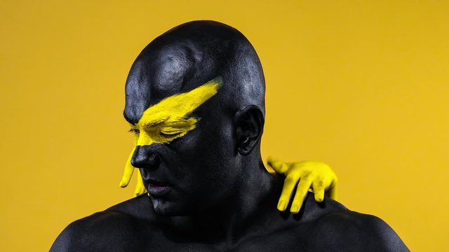 Man with female hands on the body. Bodybuilder athlete with yellow face art and black body paint. Colorful portrait of the guy with bodyart.