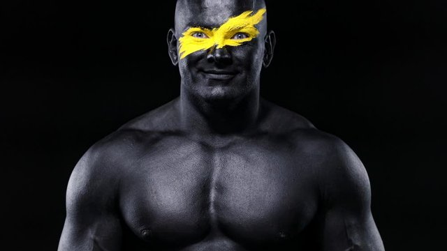 Man bodybuilder athlete with yellow color on face art and black body paint plays muscles on the chest. Colorful portrait of the guy with bodyart.