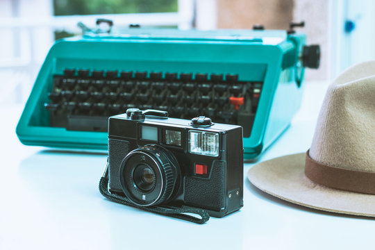 Small old photo camera with 40mm black lens next to an old green typewriter and a brown hat on a white background