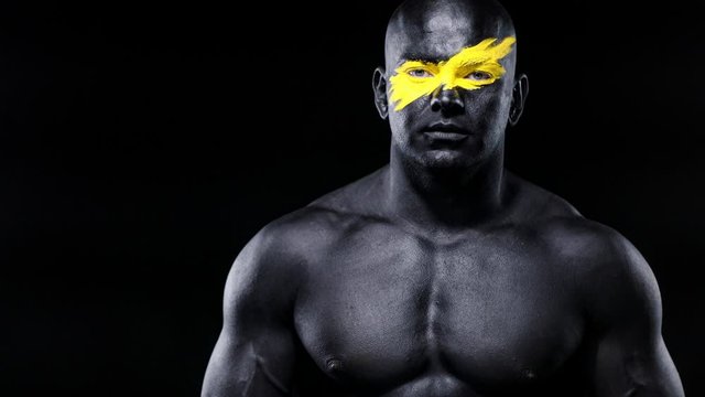 Man sports fan and bodybuilder athlete with yellow color on face art and black body paint. Colorful portrait of the guy with bodyart. Guy showing namaste sign with hands.
