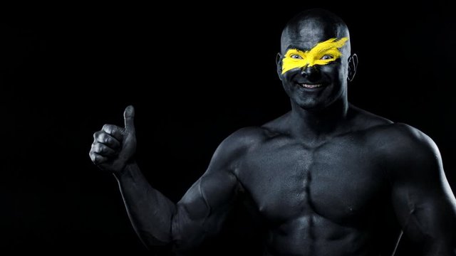Man sports fan and bodybuilder athlete with yellow color on face art and black body paint. Colorful portrait of the guy with bodyart. Guy showing thumbs up sign with fingers.