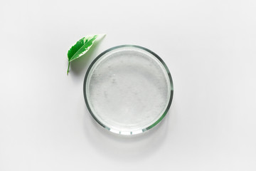 Glass petri dish with transparent pure serum for skin care and green leaf on white background, top view. Concept laboratory tests and research, making natural organic cosmetic