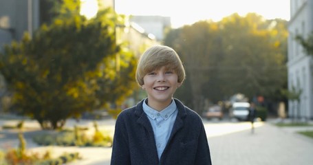 Portrait shot of cute little Caucasian boy smiling cheerfully to camera outdoors at street. Teen happy schoolboy laughing while standing outside in city on sunny summer day.