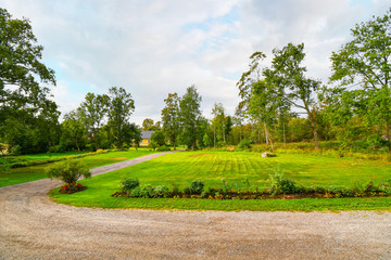 A rural dirt road runs alongside a manicured garden and lawn near a yellow secluded home in the countryside of Finland.
