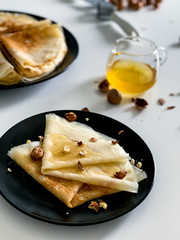 Crepes with honey and nuts, on a white plate, in black dishes. Homemade thin pancakes for breakfast or dessert.