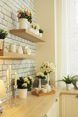 a bright cozy kitchen decorated with vases and a flower pot with artificial flowers, a wooden tabletop, shelves with decor, a floor lamp and candles, a tray with mugs and a teapot..