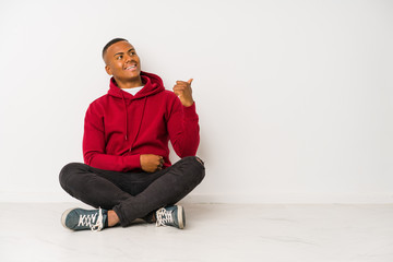 Young latin man sitting on the floor isolated points with thumb finger away, laughing and carefree.