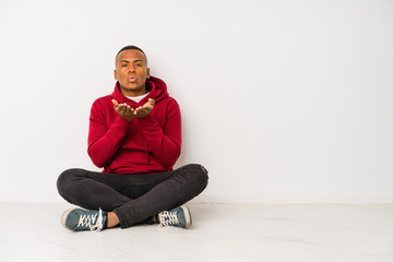 Young latin man sitting on the floor isolated folding lips and holding palms to send air kiss.