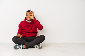 Young latin man sitting on the floor isolated excited keeping ok gesture on eye.