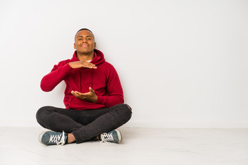 Young latin man sitting on the floor isolated holding something with both hands, product presentation.