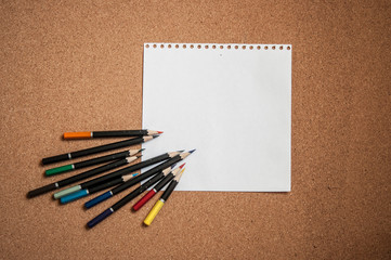 various colored pencils on cork Board background with white sheet of paper