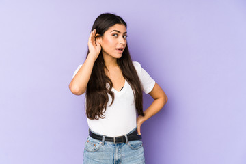 Young indian woman isolated on purple background trying to listening a gossip.