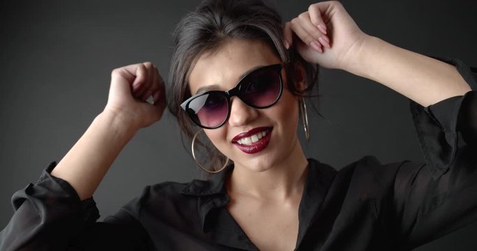 Fashion female model with curly hair wearing trendy sunglasses and sexy blouse moving actively while posing on camera. Cheerful woman isolated over dark studio background.