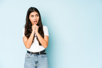 Young indian woman on blue background praying for luck, amazed and opening mouth looking to front.