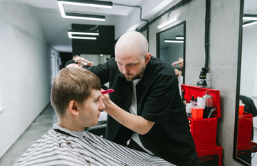 Serious male hairdresser creates a stylish hairstyle for a young client. Barber cuts hair on the head of a handsome guy. Barber shop concept.