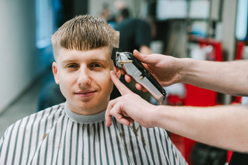 Portrait of happy barber shop customer cutting hair at professional male hairdresser, looking at camera and smiling. Barber's hand with clipper cuts the hair of a smiling young man.
