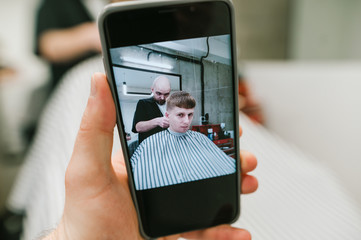 Man holds smartphone with open camera, shoots young man cuts hair in barber shop in barber shop. Smartphone takes a photo of a hairdresser trimming a handsome young man. Background