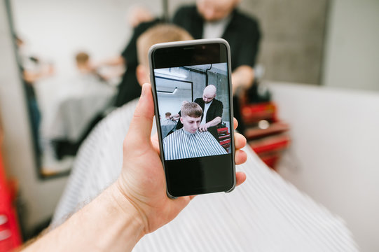 The camera on the buffer shoots a young man and a barber as he creates a stylish hairstyle. Man holds in his hand phone with camera makes photo of barber clipping client.