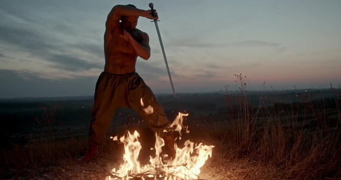 Powerful muscular shirtless man wielding a sword over the flames of a fire raising it above his head before following through with a slashing swing against a colorful sunset sky in open countryside