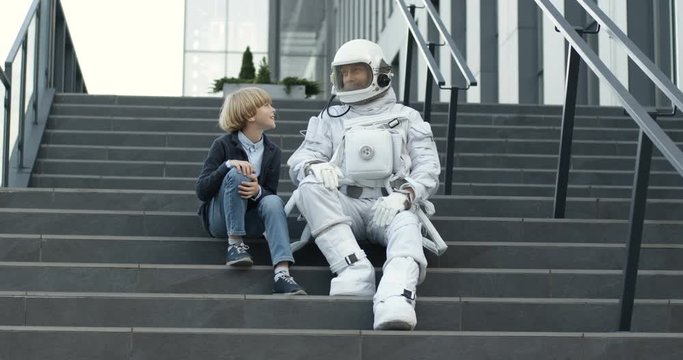 Caucasian spaceman in full armor sitting on staircase outdoors with small cute boy and telling him story about space. Little teen dreamer listening to astronaut after he coming back. Father and son.