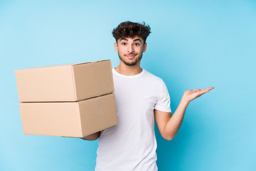 Young arab man holding boxes isolated showing a copy space on a palm and holding another hand on...