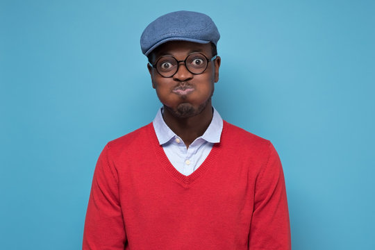 Young african man in red sweater and blue hat puffing cheeks holding breath