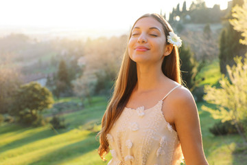 Purity and beauty concept. Portrait of beautiful girl breathing fresh air outdoor in nature with white dress and flower on ear smiling with closed eyes in spring summer time at sunset.