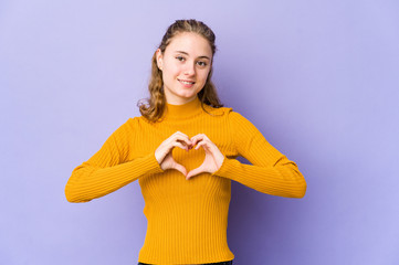Young caucasian woman on purple background smiling and showing a heart shape with hands.