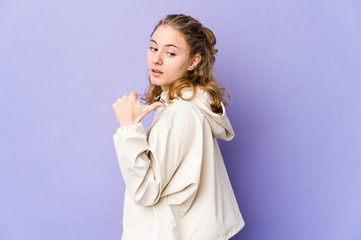 Young caucasian woman on purple background points with thumb finger away, laughing and carefree.