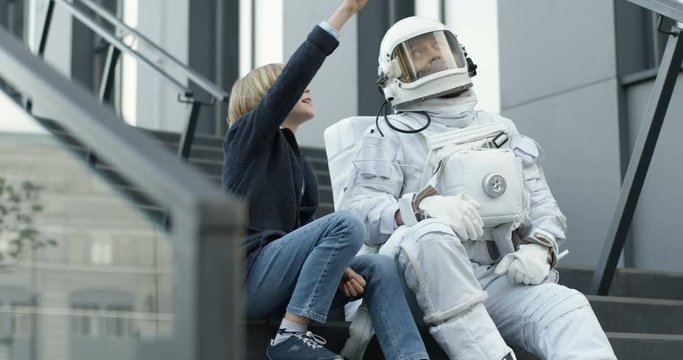 Little cute Caucasian boy sitting on staircase outdoor with spaceman in armor, talking and pointing on sky with finger. Teen small son asking father cosmonaut about cosmos.