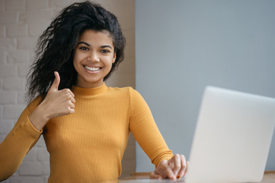 African American woman using laptop computer, showing thumb up, looking at camera and smiling, working from home. Happy student studying, learning language, watching online training courses