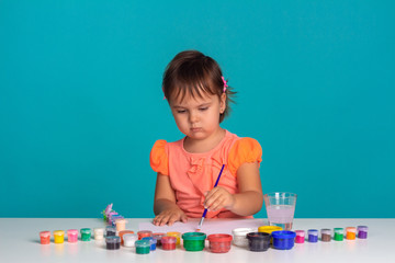 Cute little child girl drawing paints at the table. On blue background. Studio shoot.