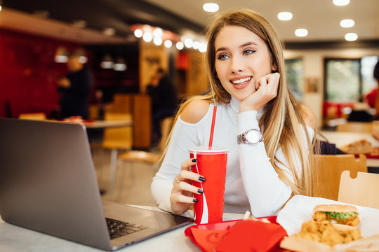 Blonde and happy girl student is learning with a laptop in a cafe, eating fast food and drinking a coca cola.