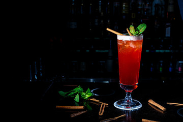 A red cocktail in a tall glass stands on the counter of a dark bar.