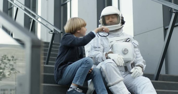Small cute Caucasian boy sitting on staircase outdoors with astronaut in space suit, talking and pointing on sky with finger. Teen little schoolboy asking spaceman about cosmos.