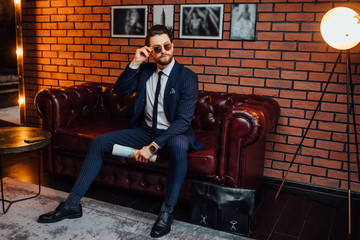 Fashionable model man holding on his eyeglasses with magazine on hands while sitting on sofa  in luxury apartament.