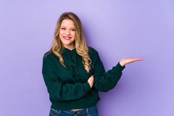 Young caucasian woman isolated on purple background showing a copy space on a palm and holding another hand on waist.