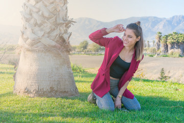 Cheerful young woman kneeling on the grass next to a palm tree with her hand on her forehead looking to the side