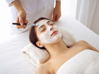 Beautiful brunette woman enjoying applying cosmetic mask with closed eyes. Relaxing treatment in medicine and spa center concepts