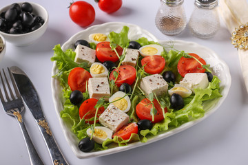 Healthy salad with cherry tomatoes, feta, quail eggs, olives and microgreens on a heart-shaped plate on a white background, healthy eating day
