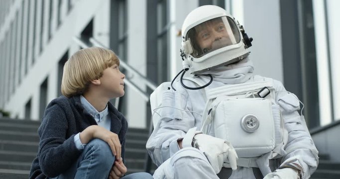 Caucasian cheerful spaceman in armor talking with cute little boy sitting on stairs outdoors and talking about space cheerfully. Small schoolboy having conversation with cosmonaut.
