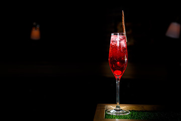 A red cocktail in a tall, narrow glass stands on the counter of a dark bar.